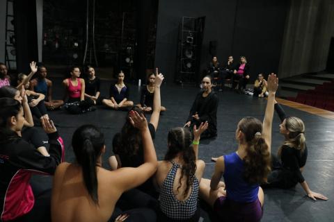 bailarinos estão sentados no chão em roda conversando. Alguns estão com as mãos para o alto como se respondendo a uma pergunta do orientador. #paratodosverem