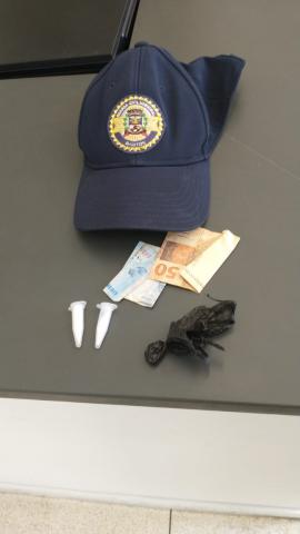 o boné da guarda municipal, sobre uma mesa, há um valor em dinheiro e duas embalagens com drogas. #paratodosverem