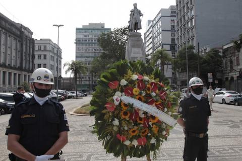 coroa de flores diante do monumento ao fundador Braz Cubas, ladeada por dois guardas municipais
