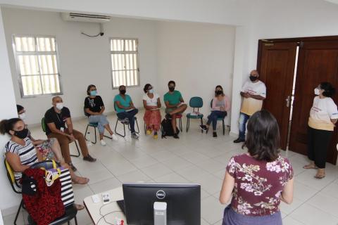 educadores estão sentados atentos a uma mulher que fala a eles. Ela está em pé e de costas para a foto. #paratodosverem