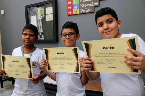três meninos posam para foto com diplomas de aluno ouvidor. #paratodosverem