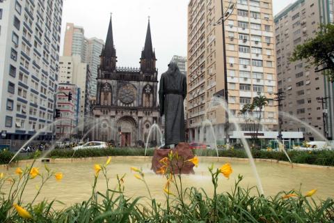 vista geral da praça diante da igreja, com santo antônio de costas para imagem e flores em primeiro plano. #paratodosverem