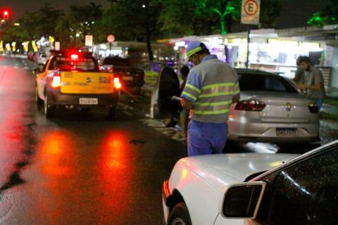 Agente da CET, com viatura à frente, está em pé na avenida. Há carros parados do lado direito da via. #paratodosverem