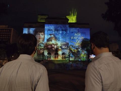 duas pessoas, em primeiro plano, observam projeção do game na fachada da prefeitura. #paratodosverem