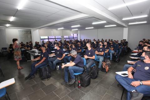 mulher está em pé falando a um grande grupo de guardas sentados em auditório. #paratodosverem