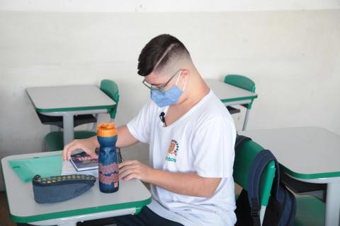 Giovanni está sentado na carteira da escola sobre a qual estão um caderno, estojo e skeeze. #paratodosverem