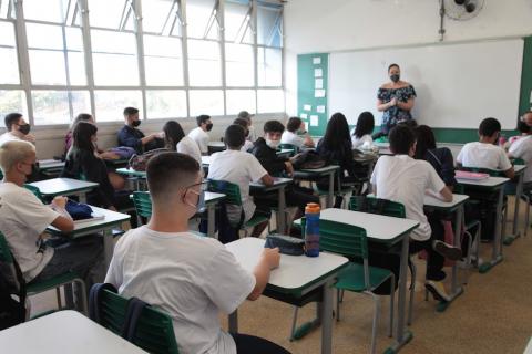 Giovanni está sentado. sala de aula está cheia. A professara está ao fundo da imagem, na frente da classe, diante de lousa. #paratodosverem
