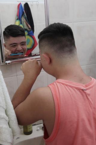 Giovanni escova os dentes na frente do espelo. Ele está de costas para a foto e seu rosto refletindo no espelho. #paratodosverem