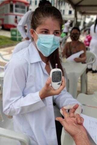mulher usando avental branco mostra aparelho de medição de glicemia. Ela esta segurando a mão de uma pessoa, de cujo dedo foi retirada gota de sangue. #paratodosverem
