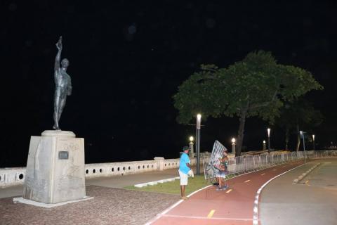 Em primeiro plano, à esquerda, o monumento em homenagem ao nadador. Ao fundo, homens instalam grades. É noite. #Paratodosverem
