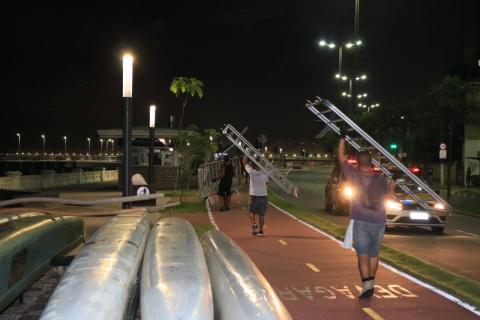 Dois homens andam pela ciclovia carregando grades. Em primeiro plano, à esquerda, canoas estacionados no solo  de cabeça para baixo. #Paratodosverem