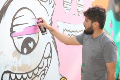 Artista aplica pintura rosa em desenho de monstro na parede. #pracegover