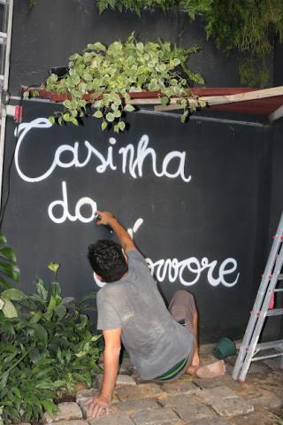 Artista, sentado, realiza pintura na área da Casinha da Àrvore. #pracegover