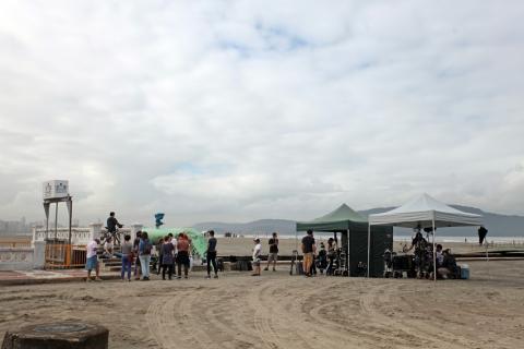 Equipe de produção está na areia da praia ao lado de barracas. #paratodosverem