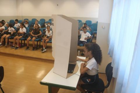 menina está dentro de cabine pronta para votar. Ao fundo, outros alunos estão sentados em auditório. #paratodosverem