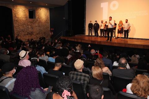 plateia atenta ao palco onde algumas pessoas estão em pé. Prefeito, à frente, fala ao público. #paratodosverem