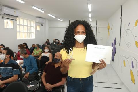 mulher posa para foto segurando o certificado na mão esquerda e uma miniatura do peixe da entrada da cidade na direita. #paratodosverem