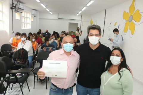 Três pessoas posam para foto. Um homem segura um certificado. #paratodosverem