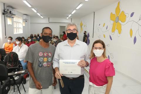 Três pessoas posam para foto. O homem, ao centro, segura um certificado. #paratodosverem