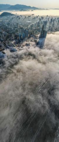 Foto aérea da cidade coberta pelo nevoeiro e prédios aparecendo ao fundo. #paratodosverem