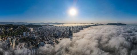 Foto aérea da cidade coberta pelo nevoeiro e prédios aparecendo ao fundo. #paratodosverem