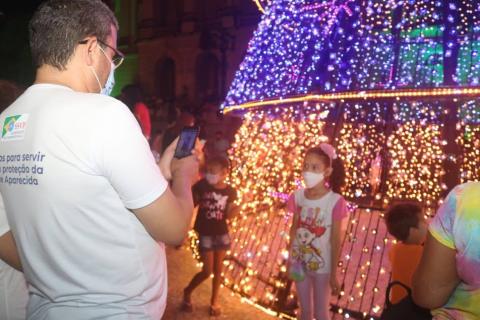 Homem fotografa menina diante de árvore iluminada. #paratodosverem
