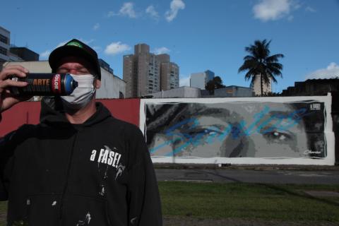 Artista coloca lata de tinta na frente do rosto com muro grafitado com Chorão ao fundo. #paratodosverem