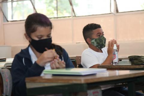 dois alunos em mesas na sala com álcool em gel #paratodosverem 