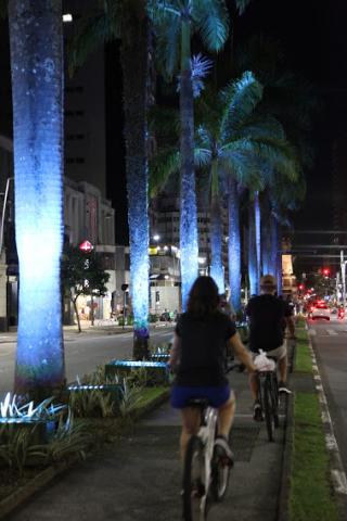 ciclistas passando por avenida iluminada de azul #paratodosverem 