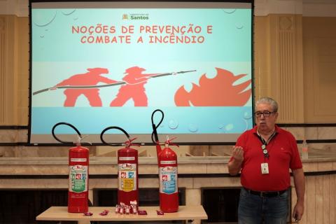 homem está em pé ao lado de uma mesa com três extintores. Ao fundo, um telão onde se lê: Noções de prevenção  e combate a incêndio. #paratodosverem