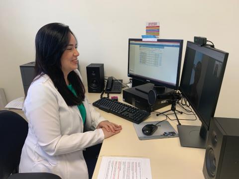 mulher usando avental branco está diante de um desktop conversando com uma pessoa que aparece no monitor. #paratodosverem