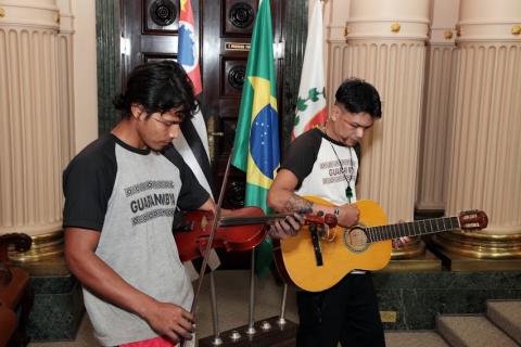 dois homens estão se apresentando. Um deles, em primeiro plano, toca violino. O outro, violão. Atrás deles estão as bandeiras do Estado, do Brasil e a de Santos. #paratodosverem