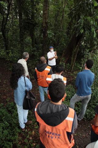 Profissionais observam técnico falando diante de uma árvore. Em primeiro plano está um homem como colete da Defesa Civil. #paratodosverem