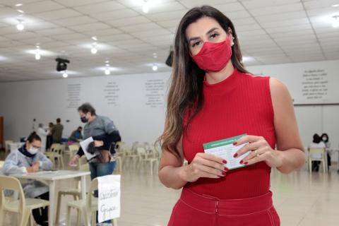 Mulher ao centro de uma sala posa para foto segurando o cartão de vacinação