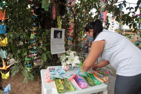 Mulher mexe em mesa branca com vários objetos coloridos e embaixo de árvore cheia de origamis. #paratodosverem