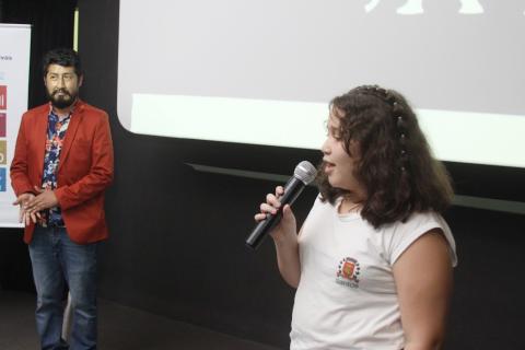 Aluna fala no microfone com o artista ao fundo da imagem. #paratodosverem