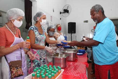 mulheres de um lado da mesa servem homem com prato na mão. #paratodosverem