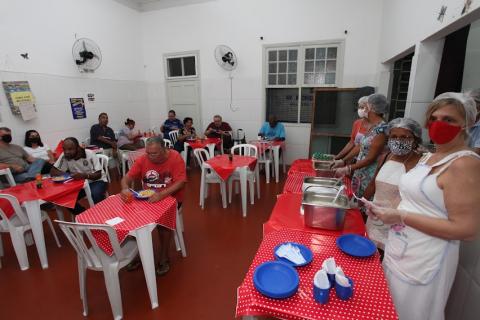 várias pessoas fazem refeição sentadas à mesa. A direita, a mesa com a comida e mulheres que  servem. #paratodosverem