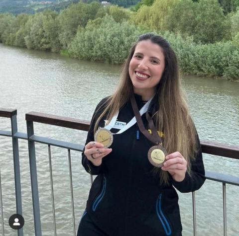 Jennifer exibe medalhas penduradas no pescoço com rio ao fundo. #paratodosverem