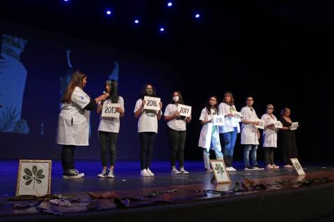 jovens estão no palco uniformizados, cada um segurando um papel com um número de ano equivalente a edições anteriores do programa. #paratodosverem