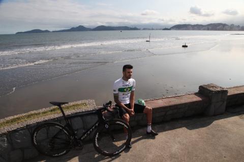 lauro sentado na mureta da praia, ao lado da bike e com a praia ao fundo. #paratodosverem
