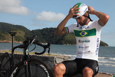 lauro está sentado em mureta da praia colocandoo capacete. A bike está ao lado. #paratodosverem