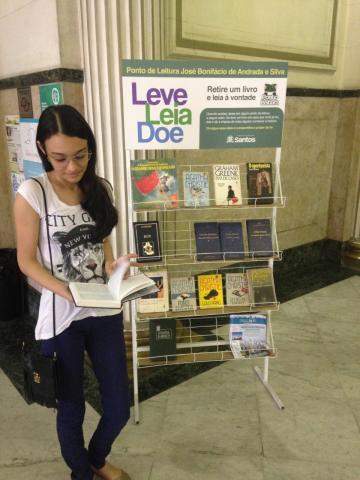 jovem está parada ao lado de estante do Leve, Leia Doa. Ela folheia uma publicação. #paratodosverem
