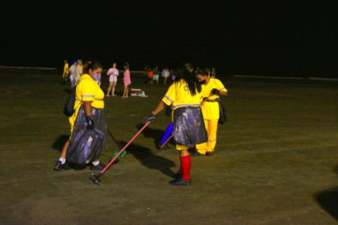 mulheres segurando sacos plásticos limpam faixa de areia.  Em primeiro plano, uma delas passa um rastelo manual na areia. #paratodosverem