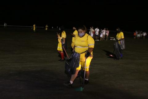 mulheres limpam faixa de areia da praia. Em pirmeiro plano, uma mulher segura saco plástico e passa o rastelo manual na areia. #paratodosverem