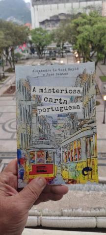 Capa do livro sendo segura por uma mão com a praça Mauá ao fundo. #paratodosverem