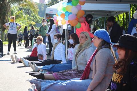 mulheres estão sentadas no chão participando de uma dinâmica. Ao fundo, tendas armadas e decoradas com balões de gás. #paratodosverem