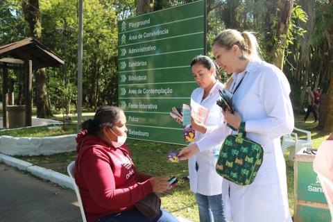 mulheres usando avental entregam folheto informativo a outra mulher que está sentada. #paratodosverem