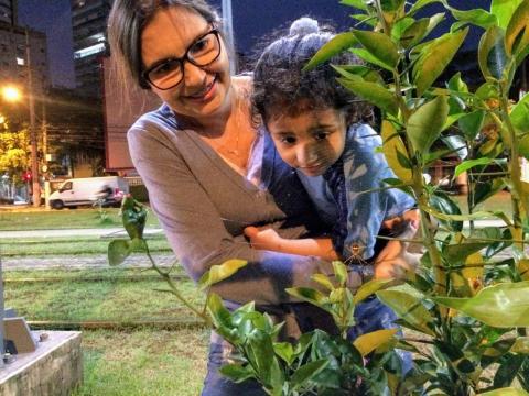 Beatriz segura a filha Manuela no colo e a aproxima de plantas. #paratodosverem