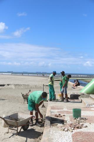 Trabalhador abaixado faz conserto em calçada próxima à faixa de areia. #pracegover